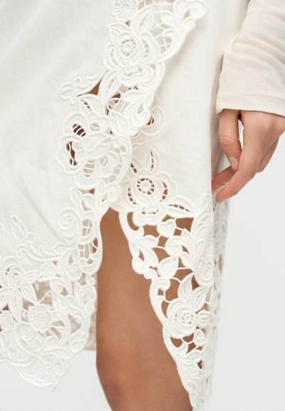 Χέρι που ακουμπάει δίπλα σε λευκό ύφασμα με περίπλοκα δαντελωτά σχέδια λουλουδιών που αποκαλύπτουν το δέρμα από κάτω.