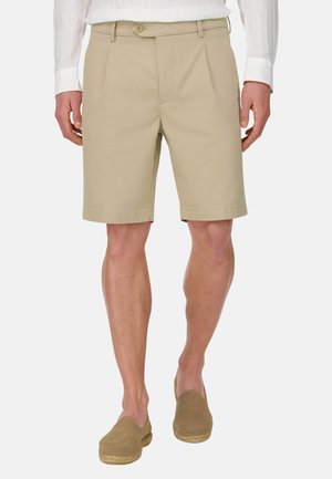 Mann trägt beige maßgeschneiderte Shorts mit Gürtelschlaufen, vorderen Falten und Knopfverschluss, kombiniert mit braunen Slip-on-Espadrilles.