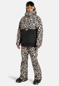 Kobieta ubrana w czarną i białą kurtkę i spodnie narciarskie w leopardzi wzór, beżową czapkę, czarne gogle narciarskie na czole oraz czarne buty śniegowe.