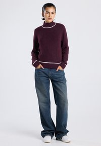 Persoon met kort haar die een bordeauxrood trui met witte rand, losse blauwe spijkerbroek en witte sneakers draagt, staand met de handen in de zakken.