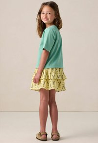 T-shirt en coton vert menthe associé à une jupe jaune superposée ornée d'un motif de homard. Des sandales marron viennent compléter la tenue.