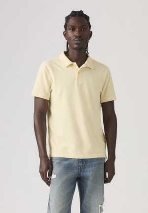 Mann mit geflochtenem Haar, der ein hellgelbes kurzärmeliges Poloshirt und zerrissene hellblaue Jeans trägt, steht vor einem einfarbigen Hintergrund.