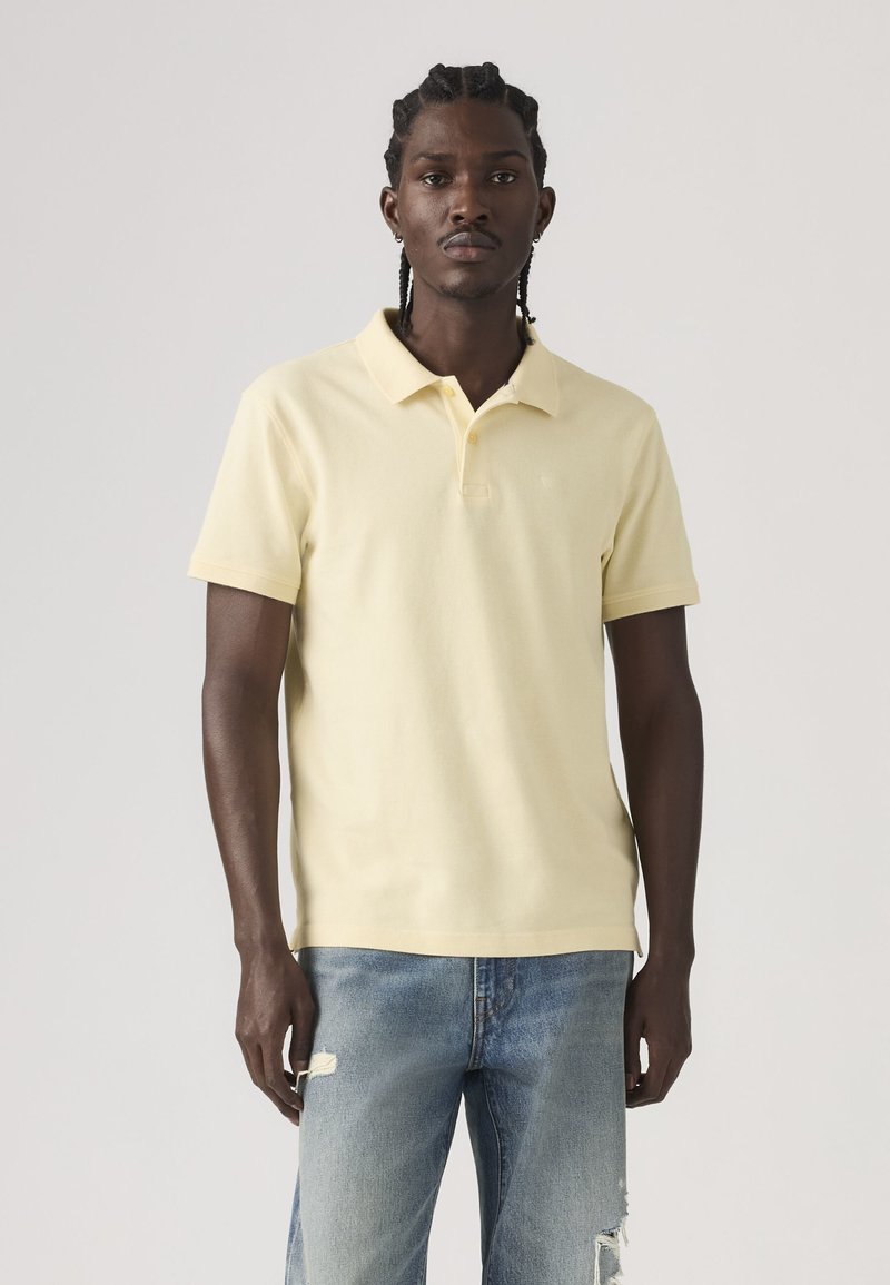 Homme aux cheveux tressés portant un polo jaune pâle à manches courtes et un jean bleu clair déchiré, debout devant un fond uni.