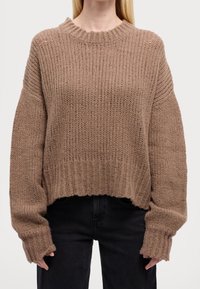 Personne portant un pull ample en tricot côtelé marron avec manches longues et un pantalon noir, montré des épaules jusqu'aux hanches.