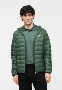 Geacă verde matlasată cu glugă, cu textură matlasată, fermoar în față și logo-ul EA7 pe pieptul stâng. Purtați peste un polo verde asortat.