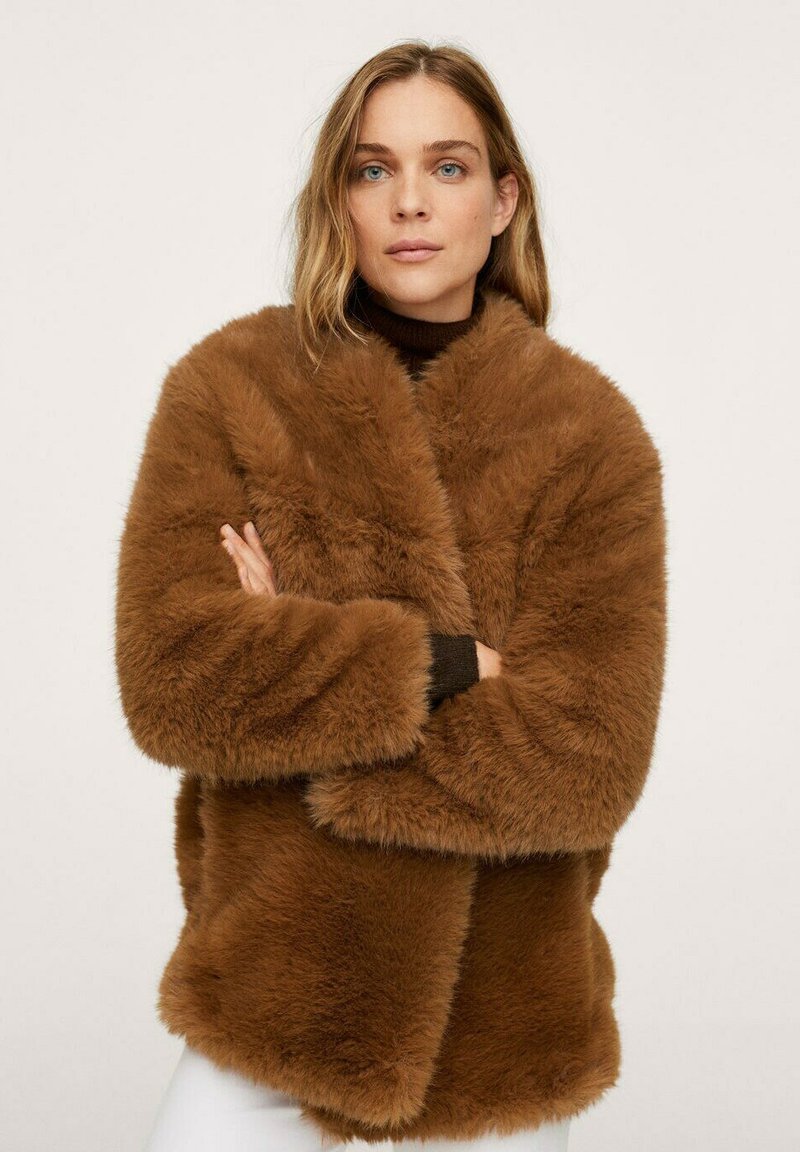 Donna con capelli castano chiaro, che indossa un cappotto in finta pelliccia marrone e un dolcevita nero, in piedi con le braccia incrociate su uno sfondo semplice.