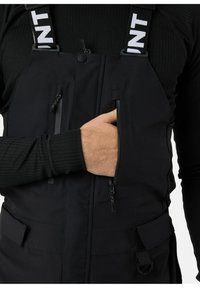 Zwarte geïsoleerde overall met verticale ritszakken, verstelbare banden en een gladde textuur. Voorzien van klittenbandaccenten en zijklussen.