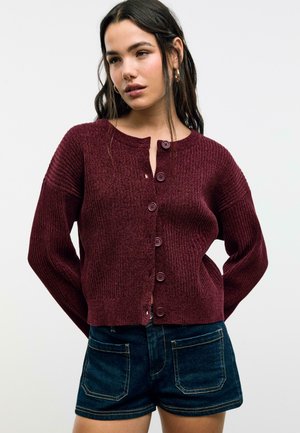 Cardigan en tricot côtelé bordeaux avec un col rond, comportant six boutons sur le devant et des manches longues. Porté sur un short en denim foncé.