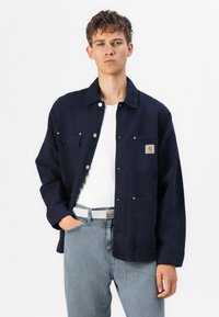 Chaqueta de mezclilla azul marino oscura con cierre de botones en el frente, dos bolsillos en el pecho y un parche con el logo de Carhartt. Lleva puesta sobre una camiseta blanca y jeans claros.