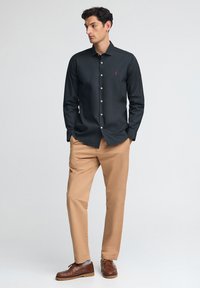 Camicia nera a maniche lunghe con bottoni bianchi e logo rosso, abbinata a pantaloni beige chiaro e scarpe marroni, in piedi contro uno sfondo neutro.