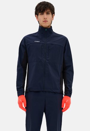 Jonge man draagt een marineblauwe jas met rits en broek met feloranje handschoenen, staande tegen een effen lichte achtergrond.