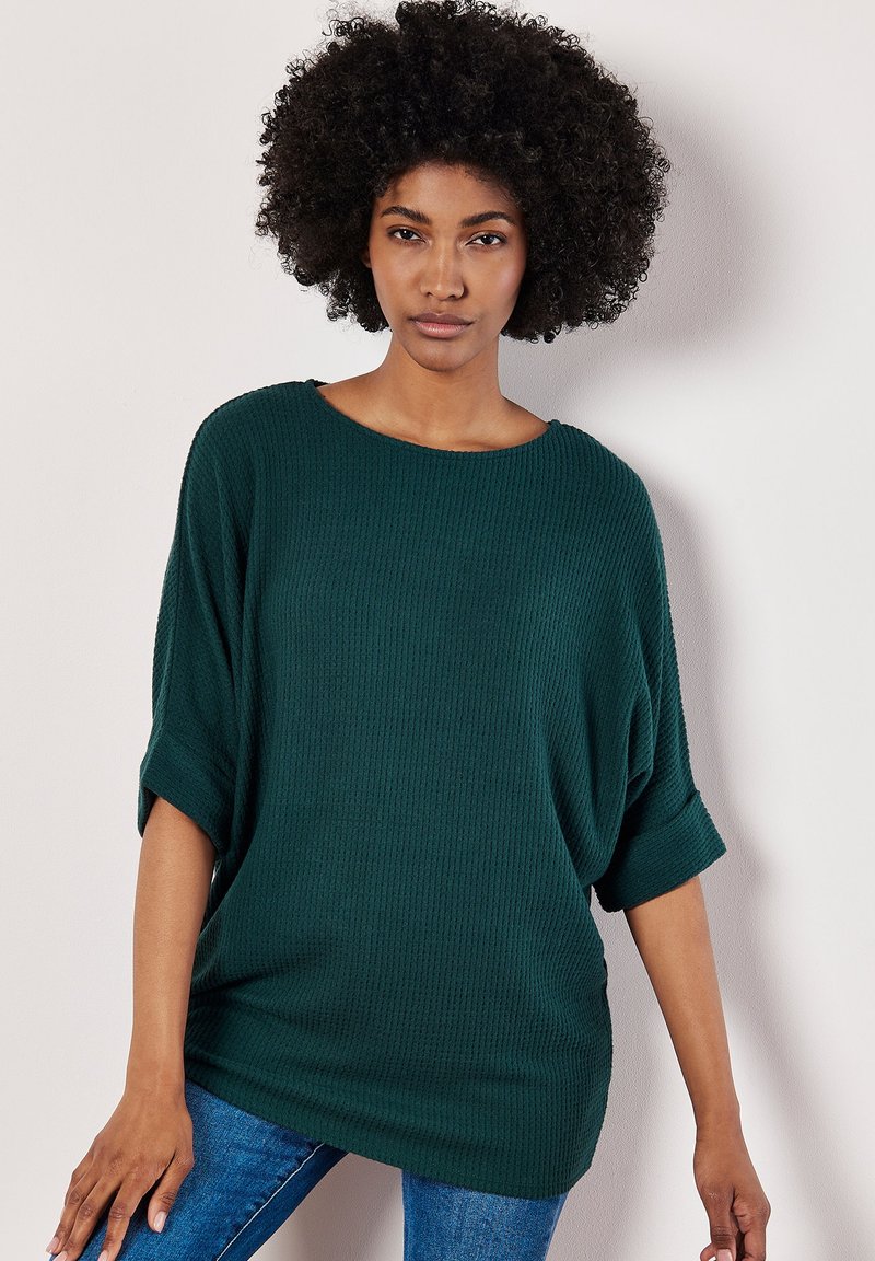 Grüner, übergroßer Waffelstrickpullover mit weitem Ausschnitt und aufgerollten Ärmeln, kombiniert mit eng anliegenden blauen Jeans. Weiche Textur, lockere Passform.