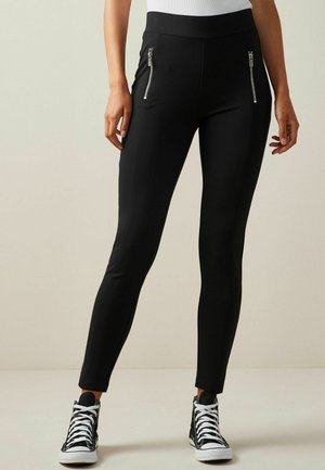 Vrouw draagt strakke zwarte broek met zilveren ritszakken aan de voorkant en zwarte hoge sneakers met witte veters en zolen.