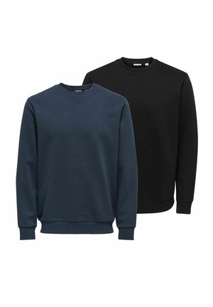Junger Mann trägt ein schwarzes Sweatshirt, lockere hellgraue Jeans und schwarze Sneaker mit niedrigem Schnitt und steht vor einem einfarbigen weißen Hintergrund.