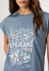 Zila īsām piedurknēm T-krekls ar baltu ziedu grafiku un tekstu "Pepe Jeans London", ar mazām rotājumiem un brīvu piegriezumu.