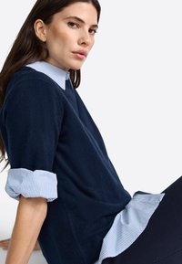 Pull bleu marine avec une finition texturée, doté de poignets de chemise rayés retroussés, superposé à une chemise à rayures bleu clair. Tissu lisse.