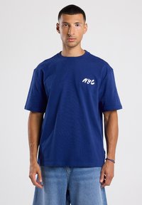 Tummansininen puuvillapaita, jossa on rento malli, lyhyet hihat ja valkoinen "NYC" -kirjaus vasemmassa rinnassa. Suora tekstuuri, rento muotoilu.