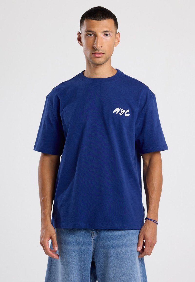 Tummansininen puuvillapaita, jossa on rento malli, lyhyet hihat ja valkoinen "NYC" -kirjaus vasemmassa rinnassa. Suora tekstuuri, rento muotoilu.