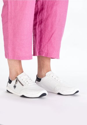 White sneakers with black accents, perforated sides, flat soles, and a zip detail. Worn with pink cropped trousers.