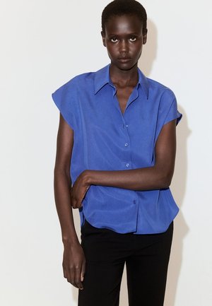 Donna dalla pelle scura che indossa una camicia blu a maniche corte con bottoni e pantaloni neri, in piedi davanti a uno sfondo chiaro e uniforme.