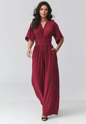 Kobieta z długimi kręconymi włosami, ubrana w płynący bordowy kombinezon z szerokimi nogawkami i krótkimi rękawami, stojąca na jednolitym tle.