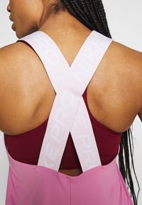 Top deportivo Nike Pro que presenta un diseño entrelazado con tirantes de color rosa claro sobre una base de color burdeos profundo, fabricado con un tejido suave y transpirable.