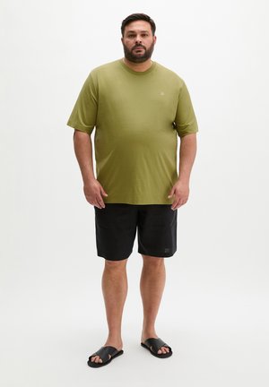 Man staat tegen een effen achtergrond, draagt een olijfgroen T-shirt, zwarte korte broek en zwarte open-teen slippers, kijkt met een neutrale uitdrukking naar voren.