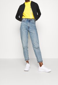 Chaqueta negra con cremallera, camiseta amarilla con estampado gráfico, vaqueros de tiro alto azul claro y zapatillas deportivas blancas. Atuendo casual.