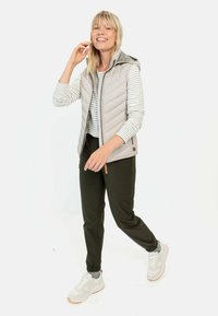 Leichte, gesteppte beige Weste mit Kapuze, kombiniert mit einem gestreiften Langarmshirt und dunkelgrünen Hosen. Legere Sneakers mit Akzenten.