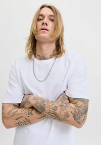 Balta medvilninė marškinėliai su trumpomis rankovėmis, apvaliu kaklu ir mažu logo. Asmuo turi ilgus plaukus, grandinėlę ant kaklo ir tatuiruotas rankas.