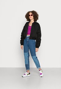 Casaco bomber preto, cropped roxo, jeans de patchwork em tons de azul, e ténis brancos com detalhes em rosa e verde. Cabelo encaracolado, óculos de sol redondos cor-de-rosa.