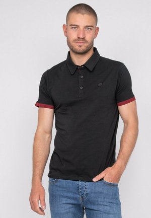 Homme aux cheveux courts et barbe portant un polo noir avec des bords de manches rouges et un jean bleu, debout avec une main dans la poche.