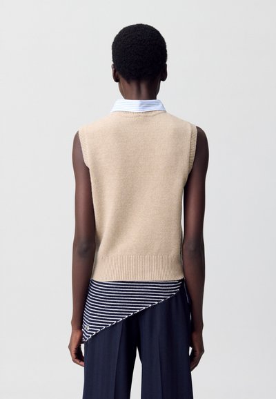 Maglione beige senza maniche realizzato in maglia con orlo a coste, sovrapposto a un tessuto a righe blu navy e bianco, creando un design asimmetrico.