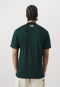 Mies, jolla on letitetyt hiukset, pukee ylleen tummanvihreän lyhythihaisen paidan, jossa on pieni värikäs logo yläselässä, sekä vaaleanbeiget housut.