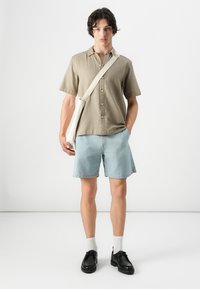 Chemise beige clair à manches courtes avec fermeture à boutons, associée à un short en denim bleu clair et des chaussures noires. Sac à bandoulière blanc visible.