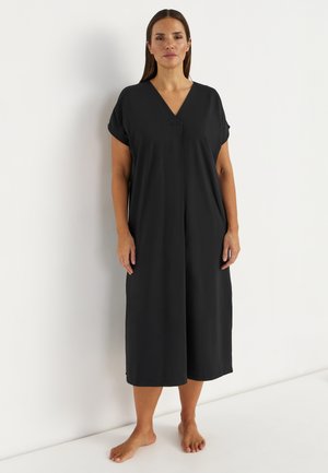 Kobieta stojąca boso, ubrana w luźną, czarną sukienkę midi z krótkim rękawem i dekoltem w serek na jednolitym białym tle.