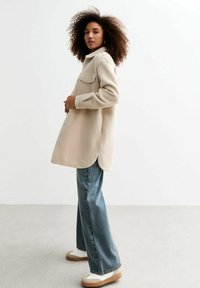 Abrigo de mezcla de lana beige con cuello, bolsillos laterales visibles y un corte holgado, combinado con jeans de pierna ancha de color azul claro y zapatillas beige.