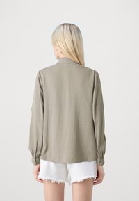 Kvinna med långt blont hår som bär en lös, långärmad olivfärgad skjorta och vita fransade denimshorts, stående mot en enkel ljus bakgrund.