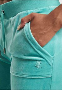 Turkusowe dresy o miękkiej fakturze, z elastycznym paskiem w talii, boczną kieszenią i haftowanym logo. Posiadają regulację sznurkiem.