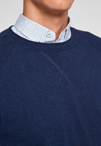 Homme portant un pull bleu à col rond par-dessus une chemise blanche boutonnée avec des motifs bleus, vu du cou à la poitrine.