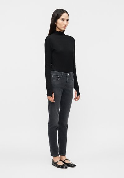 Jeune femme aux longs cheveux foncés portant un pull à col roulé noir à manches longues, un jean gris foncé et des chaussures Mary Jane noires, debout devant un fond blanc.