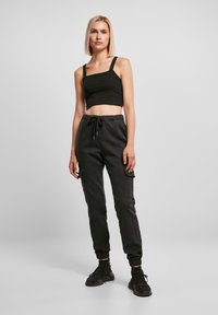 Žena s blond bob účesom, oblečená v čiernom strečovom topi, čiernych joggerových nohaviciach s viažucou šnúrkou a čiernych teniskách na jednofarebnom pozadí.
