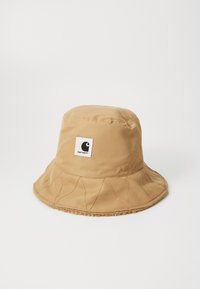 Chapeau seau en tissu beige avec une couronne texturée, un large bord et des coutures contrastées. Présente un patch logo carré sur le devant.