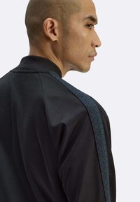 Giacca sportiva nera con colletto a coste, caratterizzata da una striscia verde testurizzata lungo la spalla. Tessuto liscio con dettagli minimi.