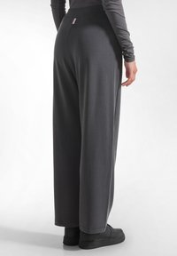 Grijze, losse broek gemaakt van zachte stof. Heeft een elastische tailleband en wijde pijpen. Zwarte schoenen zijn zichtbaar aan de voeten van het model.