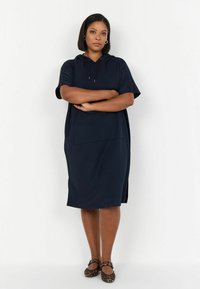 Vestido azul marino con mangas cortas, capucha y bolsillo delantero. El material es suave con un ajuste relajado, y cuenta con aberturas laterales para facilitar el movimiento.