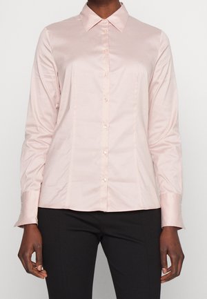 Woman wearing a light pink satin button-up long-sleeve shirt and black pants, standing against a plain white background.