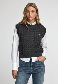 Grå stickad ärmlös tröja med ett kabelmönster, ribbad nederkant och rundad hals, över en vit skjorta med krage. Jeanjacka synlig.