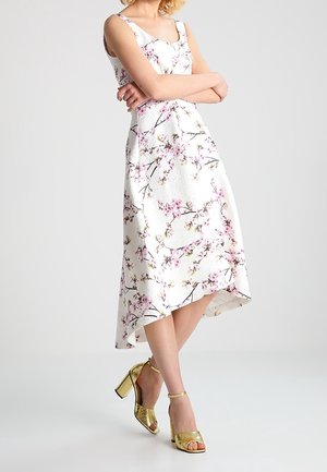 Bloemen midi-jurk in het wit met roze kersenbloesem patronen, voorzien van dunne bandjes en een high-low zoom. Gouden sandalen met blokhak.