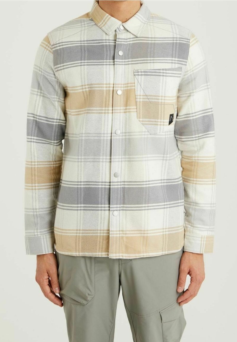 Homme portant une chemise à carreaux beige, grise et blanche boutonnée avec une poche poitrine et un pantalon cargo gris clair, debout devant un fond uni.